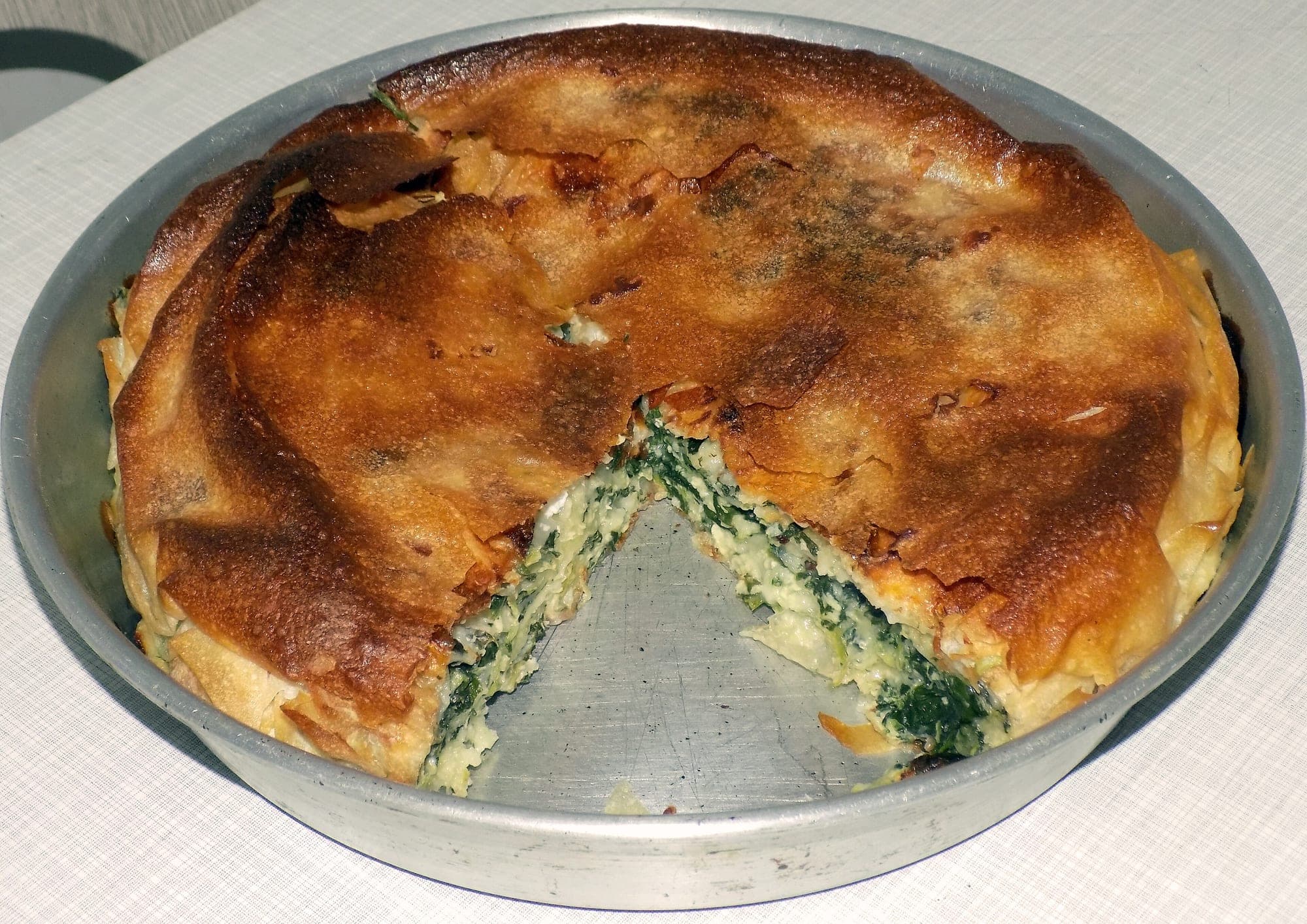 Homemade spinach and cheese byrek in a round baking pan, with one slice cut to show the green filling between golden layers of pastry